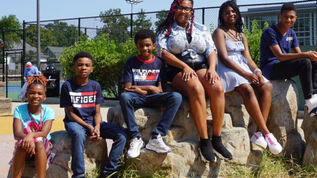 Mother with five pre-teen and teen children sitting on rock formation at playground smiling for the camera.