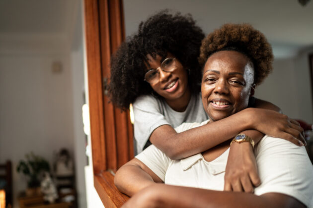 Black grandmother and teen granddaughter smiling, posed with granddaughter's arms around grandmother.