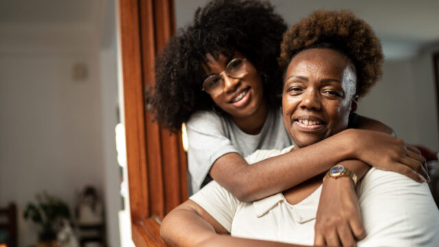 Black grandmother and teen granddaughter smiling, posed with granddaughter's arms around grandmother.