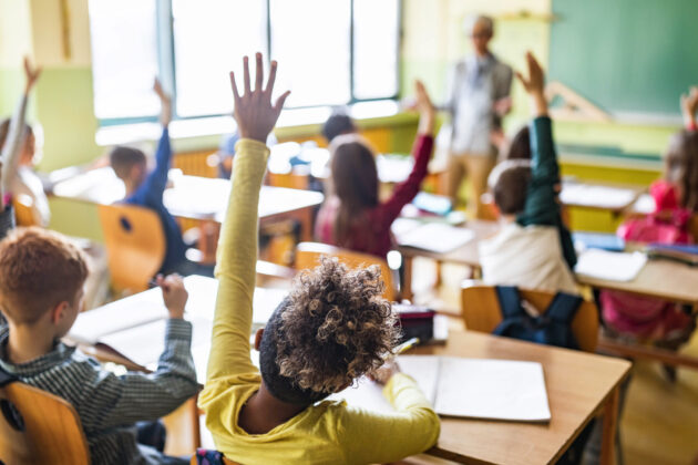 Photo of elementary school classroom with many kids seated at desks, hands raised.