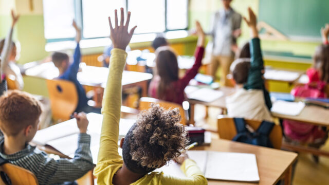 Photo of elementary school classroom with many kids seated at desks, hands raised.