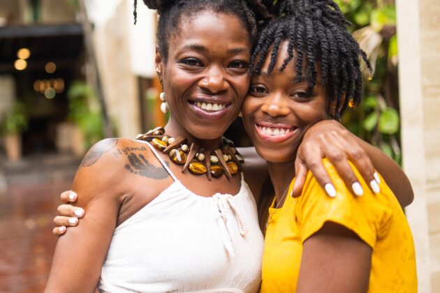 Portrait photo outdoors of mother and daughter smiling at camera with arms around each other's shoulders.