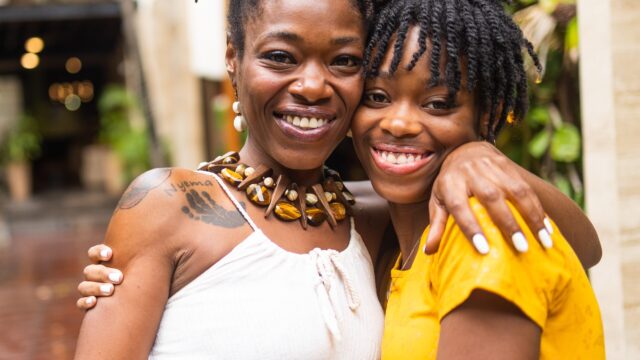 Portrait photo outdoors of mother and daughter smiling at camera with arms around each other's shoulders.