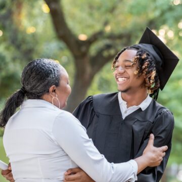 Photo of woman in white shirt embracing smiling young man in black graduation robe and mortarboard. They are outside, and a tree is visible in the background above them.