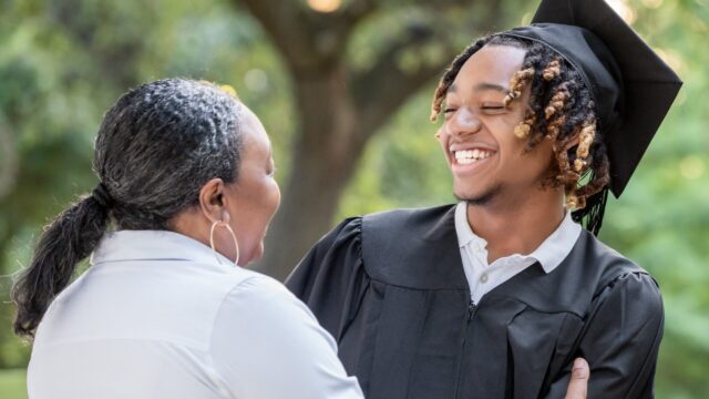 Photo of woman in white shirt embracing smiling young man in black graduation robe and mortarboard. They are outside, and a tree is visible in the background above them.