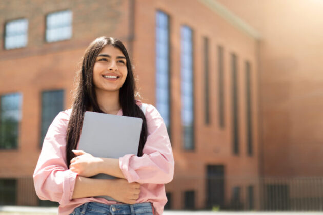 Young woman holding laptop in front of school building.