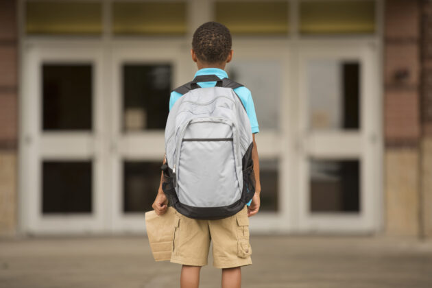 Photo of the back of a young student with a large backpack, holding a brown bag lunch, looking ahead at the doors to school.