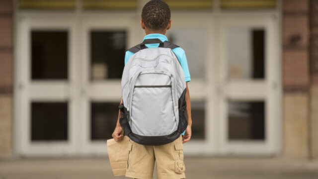Photo of the back of a young student with a large backpack, holding a brown bag lunch, looking ahead at the doors to school.