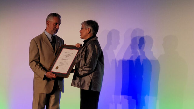 Chad Sarchio presents award plaque to Judith Sandalow with stage lighting casting their shadows in blue on the screen behind them.